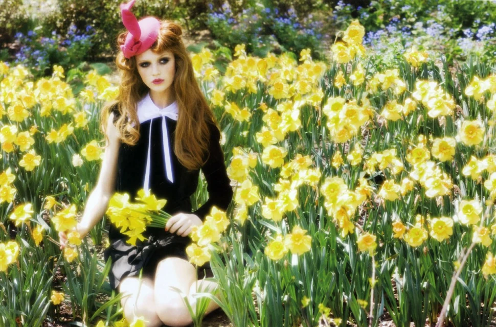 Young woman in a black dress and pink hat sits among vibrant yellow daffodils in a sunny garden setting, holding a bunch of flowers.