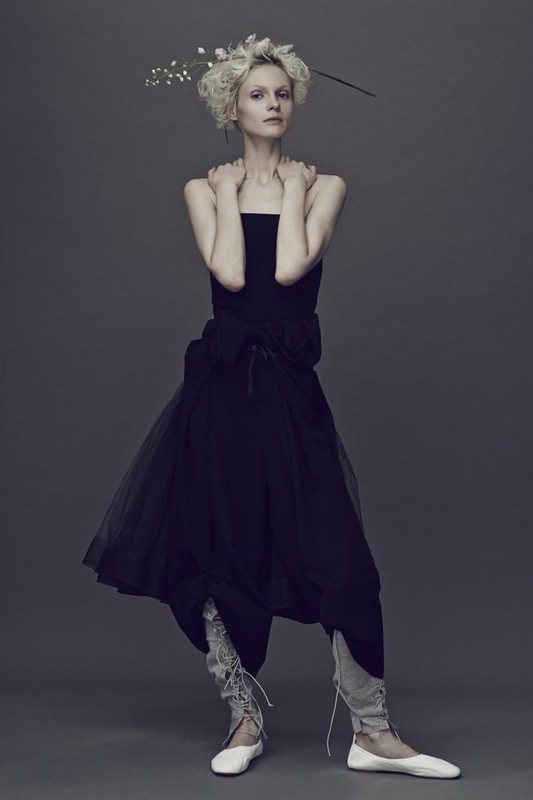 A slender woman poses in a strapless black dress with a tulle skirt, paired with leggings and ballet flats, against a grey background.