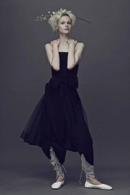 A slender woman poses in a strapless black dress with a tulle skirt, paired with leggings and ballet flats, against a grey background.