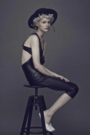 Model with blonde hair and black hat seated on stool wearing black leather jumpsuit and white shoes.