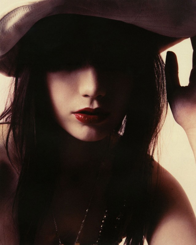 Woman with dark hair poses with a white hat and red lipstick. The image features dark shadows and a focus on her face and jewelry.