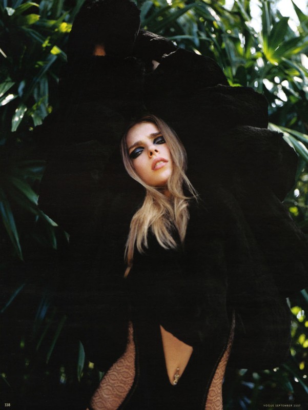 Young woman in a black dress and dark eye makeup poses in a jungle-like environment. The lighting is bright, casting a glow on her face.
