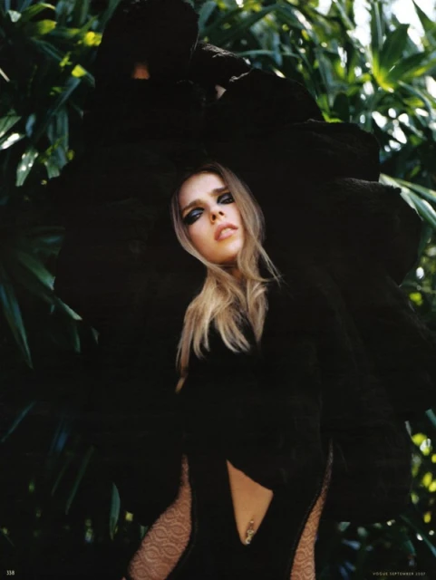 Young woman in a black dress and dark eye makeup poses in a jungle-like environment. The lighting is bright, casting a glow on her face.