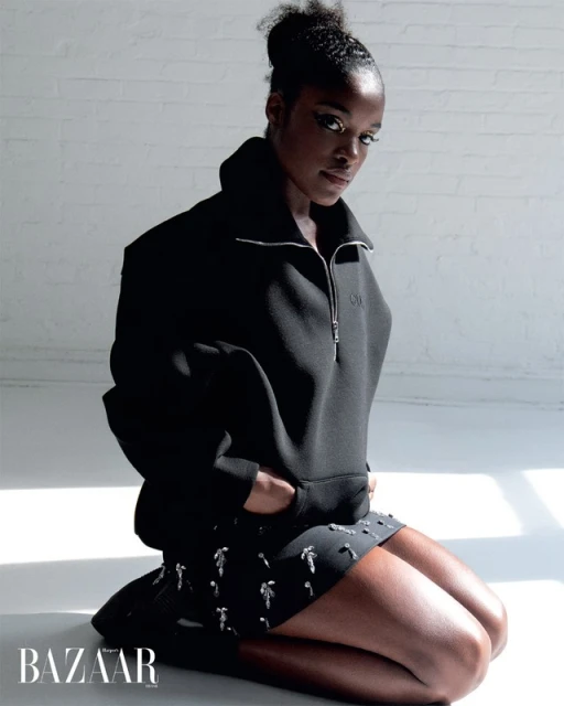 Portrait of an African American woman with dark hair and wearing a black pullover and embellished black skirt, sitting on floor in a studio.