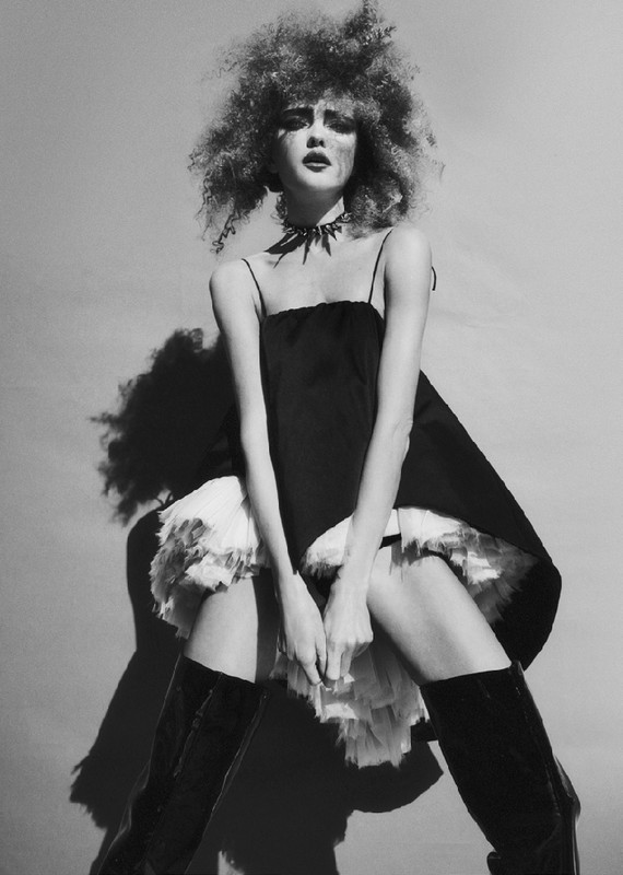 Monochrome fashion portrait of a young woman with an afro hairstyle wearing a short black dress and high boots against a grey backdrop.