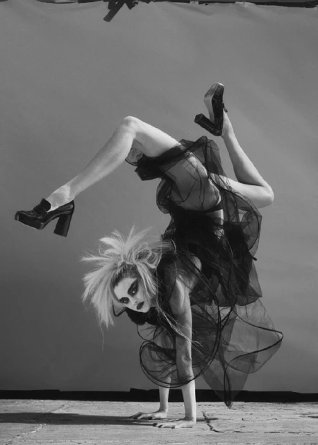 Woman with dramatic makeup performs a handstand wearing black tulle and platform heels in a studio with a grey background and concrete floor.