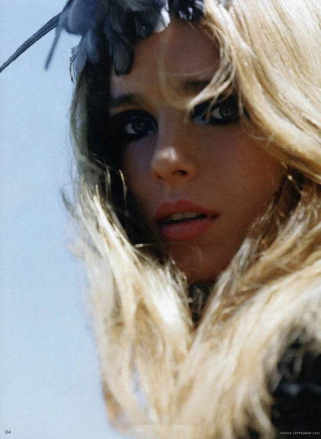Glamour shot of a blonde woman with heavy dark eye makeup and a feather headpiece, close-up in outdoor setting with a light blue sky.