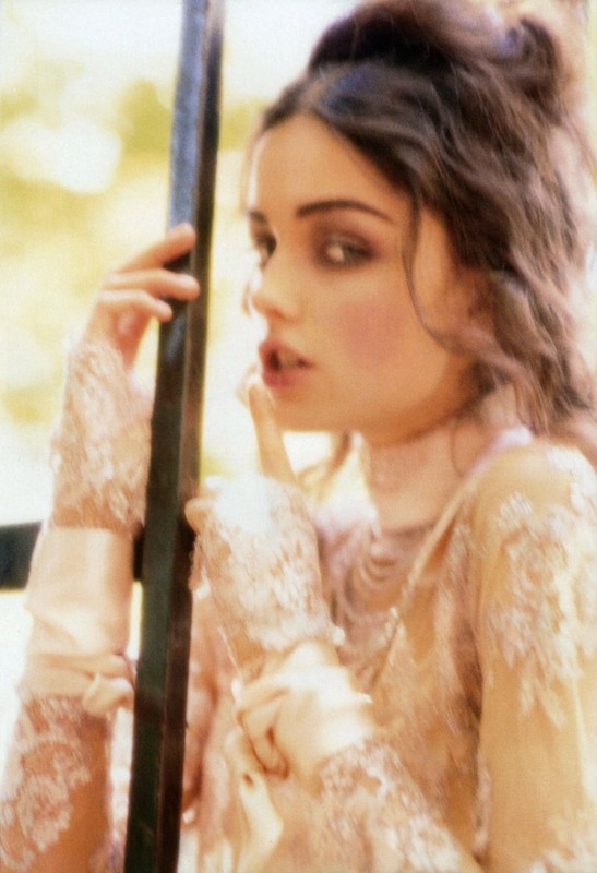 Woman in a lace top poses, holding a finger to her lips. She has dark hair pulled back with soft makeup in an outdoor environment.