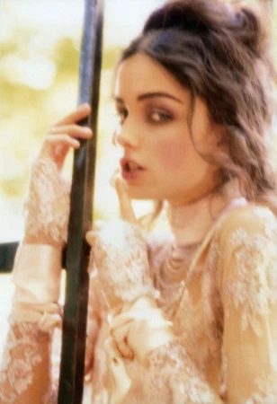 Woman in a lace top poses, holding a finger to her lips. She has dark hair pulled back with soft makeup in an outdoor environment.