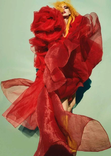 Fashion shot of a person with yellow hair wearing a large, voluminous red dress with ruffles, set against a light-green backdrop.