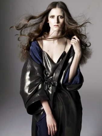 Model with long blowing hair wearing a metallic coat over a blue dress against a grey backdrop