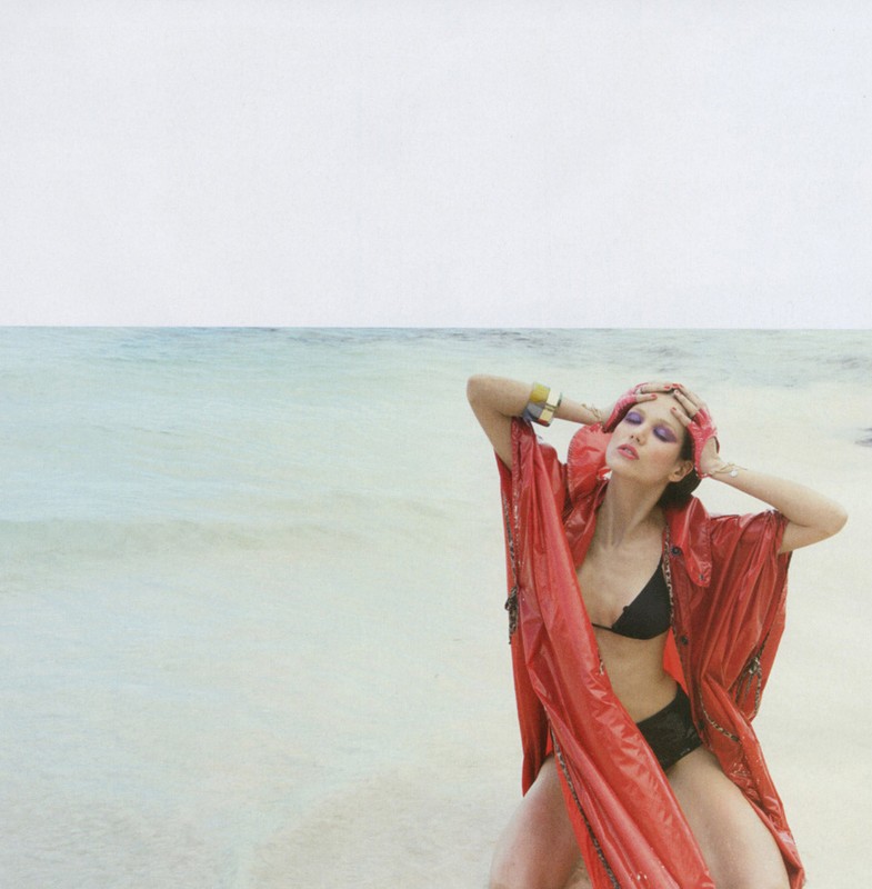 Woman poses in red robe and black bikini on a beach with ocean in the background, accessorized with gold cuffs and forehead jewelry.