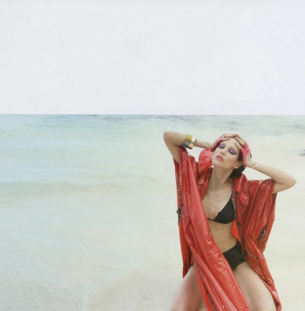 Woman poses in red robe and black bikini on a beach with ocean in the background, accessorized with gold cuffs and forehead jewelry.