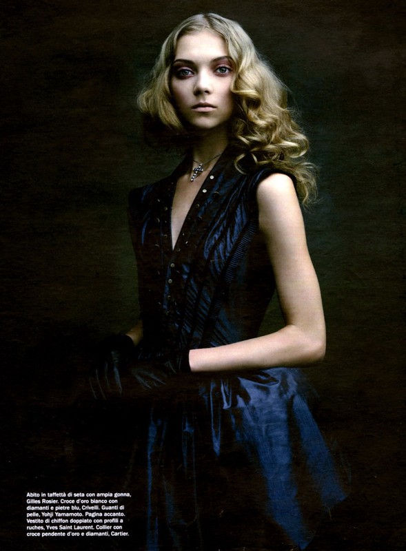 Portrait of a blonde woman wearing a dark sleeveless dress and pendant necklace against a moody textured background.
