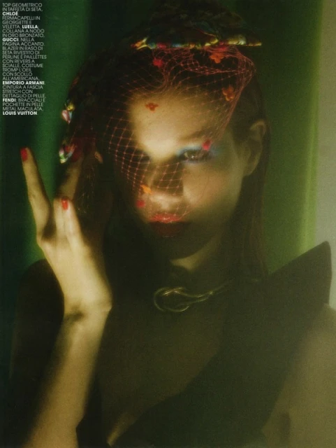 Woman with red nails and colorful eye makeup wearing a black dress and fishnet veil in a studio setting with green background.