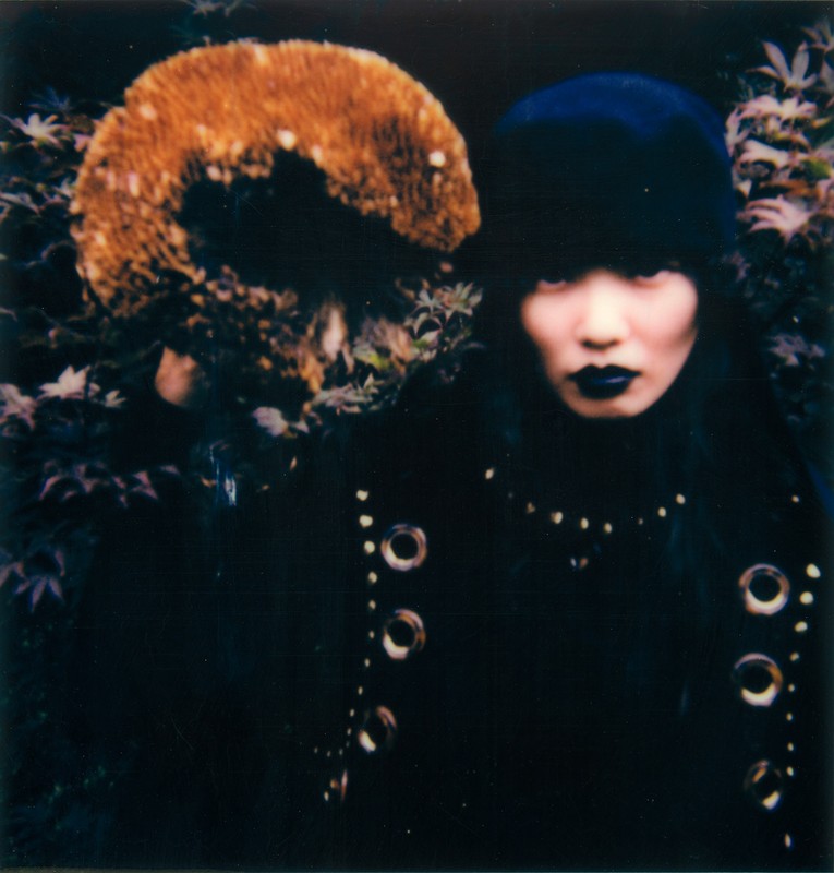Asian woman in dark coat with metal details holds large mushroom with textured cap amid foliage in a close-up, moody outdoor shot.