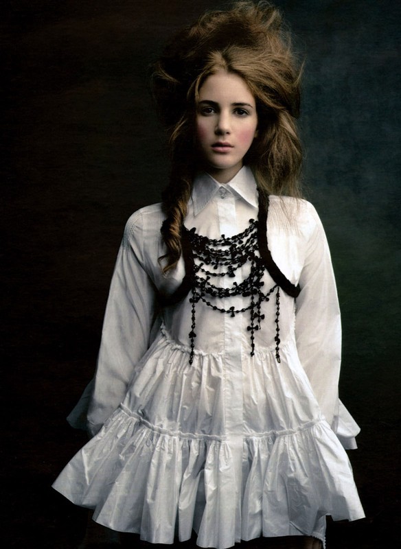 A young woman with brown hair and pale skin wears a white frilly dress and beaded necklace against a dark, moody background.