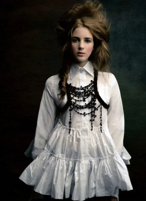 A young woman with brown hair and pale skin wears a white frilly dress and beaded necklace against a dark, moody background.