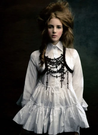A young woman with brown hair and pale skin wears a white frilly dress and beaded necklace against a dark, moody background.