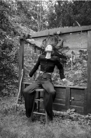 Monochrome portrait of a woman with long hair tossed back, seated on a wooden stool outdoors with garden structures.