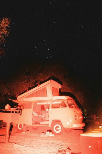 Vintage camper van with a pop-up roof parked outdoors at night, illuminated by a warm light source, with a starry sky visible above.