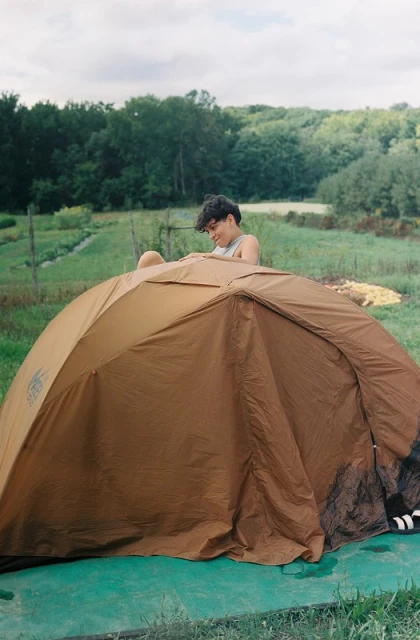 Young adult woman setting up a brown camping tent on green grass in a yard, with a green forest visible in the background.
