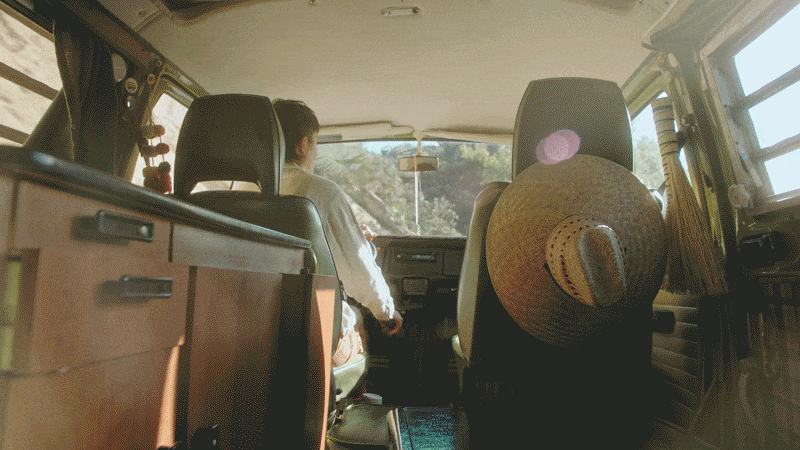 Inside a vintage VW van, a person drives through a sunny, wooded area, evoking a sense of travel and adventure with retro charm.