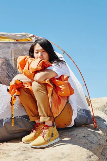 An Asian male sits on a rock with a white tent behind him and wearing orange jacket, tan pants, and tan hiking boots, beneath a blue sky.