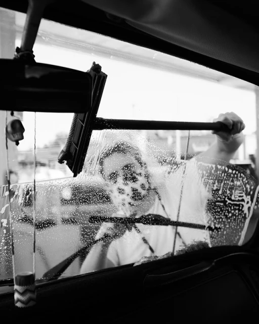 Black and white shot of a man cleaning a car windshield with a squeegee, creating a soapy texture over the glass.