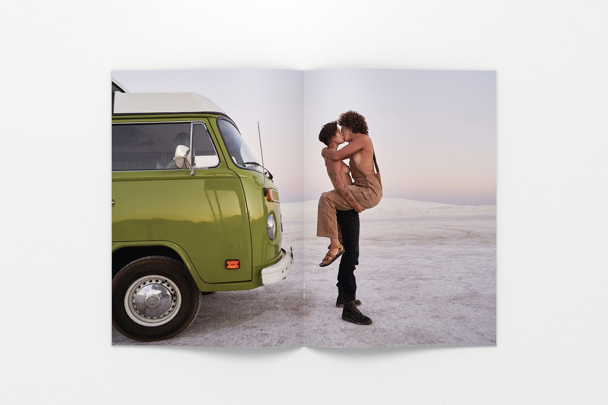 A couple embraces near a vintage green Volkswagen van against a backdrop of a light sky and white sandy terrain.
