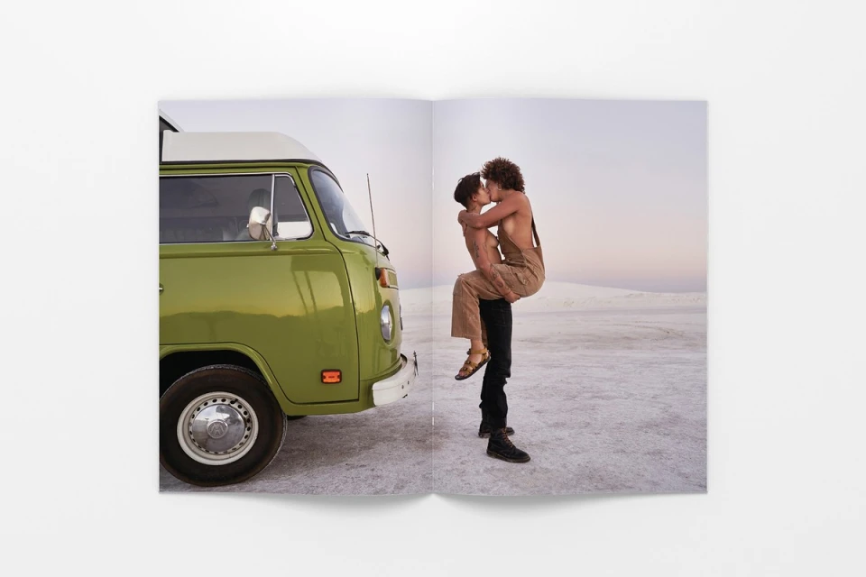 A couple embraces near a vintage green Volkswagen van against a backdrop of a light sky and white sandy terrain.