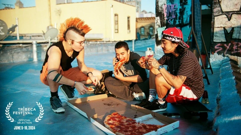 Three young adults with alternative hairstyles and casual clothing are sitting on a rooftop eating pizza from a cardboard box.