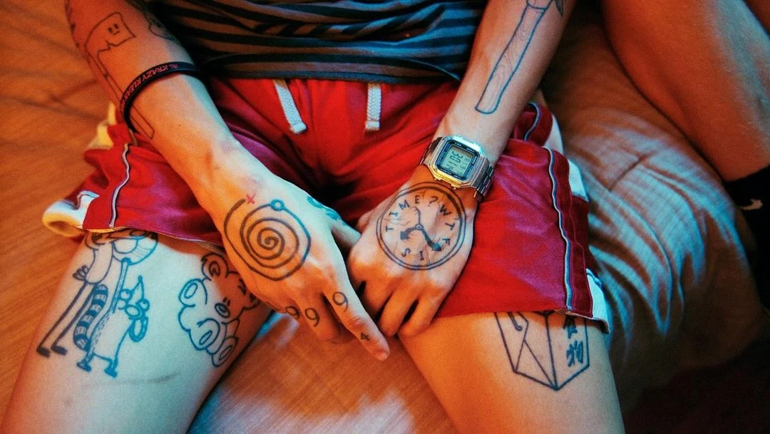 Close-up of person with tattoos on arms, hands, and legs, wearing a striped top, shorts and a digital watch.