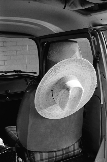 Wide-brimmed straw hat rests on a car seat inside an automobile, illuminated in grayscale. Vintage car interior visible.