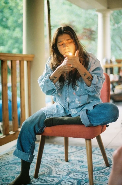 A young woman sits on a chair, indoors, lighting a cigarette with a lighter, wearing denim shirt and pants with dark socks.