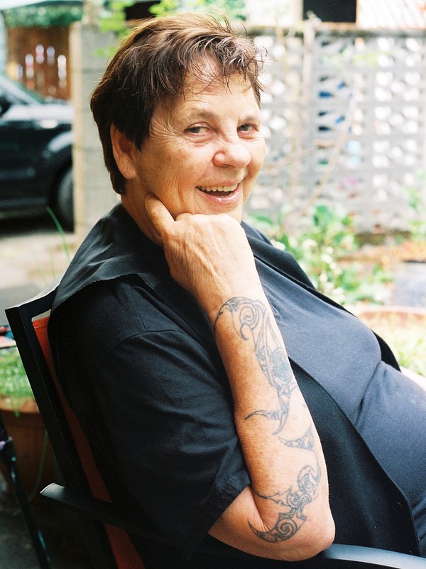 Smiling mature woman with arm tattoo seated outdoors in a black shirt with her hand to her face in front of greenery.