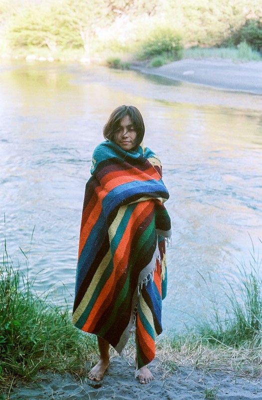 A young adult stands wrapped in a striped blanket near a river, with greenery and trees visible in the background on an overcast day.
