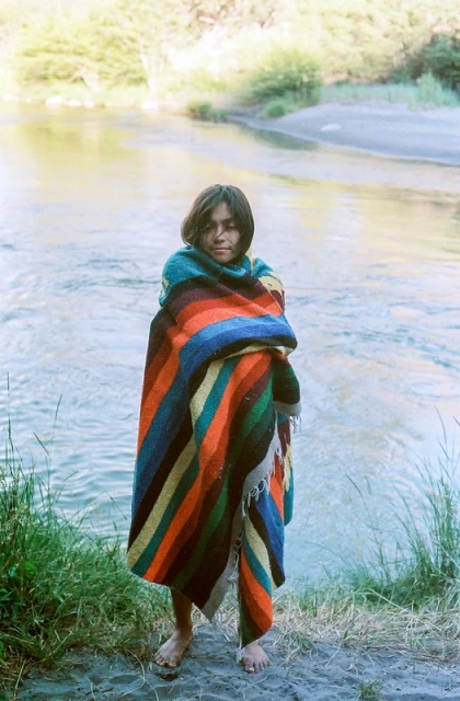 A young adult stands wrapped in a striped blanket near a river, with greenery and trees visible in the background on an overcast day.