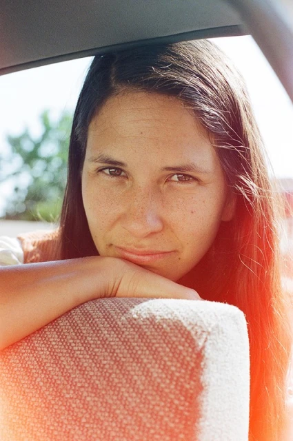 Close-up portrait of a woman resting her chin on her hand in a car, with brown hair and eyes, fair skin, and warm, diffused natural light.