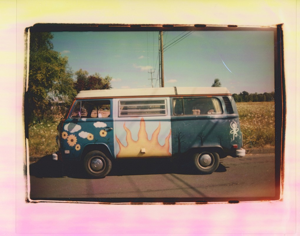 Decorated vintage Volkswagen van parked on the roadside, featuring sun, cloud, and tree art, with a field and blue sky backdrop.