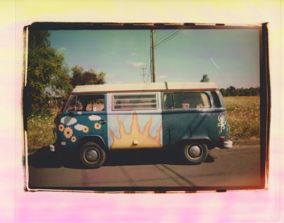 Decorated vintage Volkswagen van parked on the roadside, featuring sun, cloud, and tree art, with a field and blue sky backdrop.