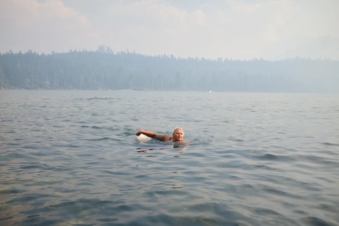 A person swimming in a vast, calm lake under a hazy sky with distant tree-lined shores