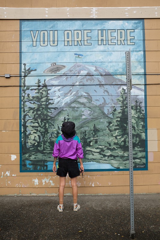 Person in a purple jacket looks at a mural of a mountain with a UFO and text reading, 'You Are Here,' on the side of a building.