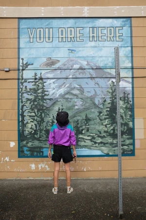 Person in a purple jacket looks at a mural of a mountain with a UFO and text reading, 'You Are Here,' on the side of a building.