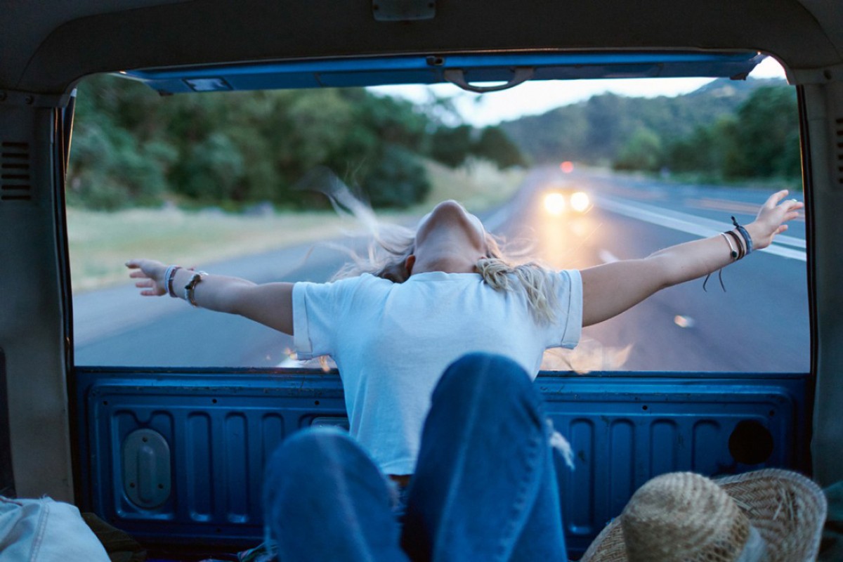View from inside vehicle of female wearing blue jeans with arms open looking at road, trees and distant car headlights.