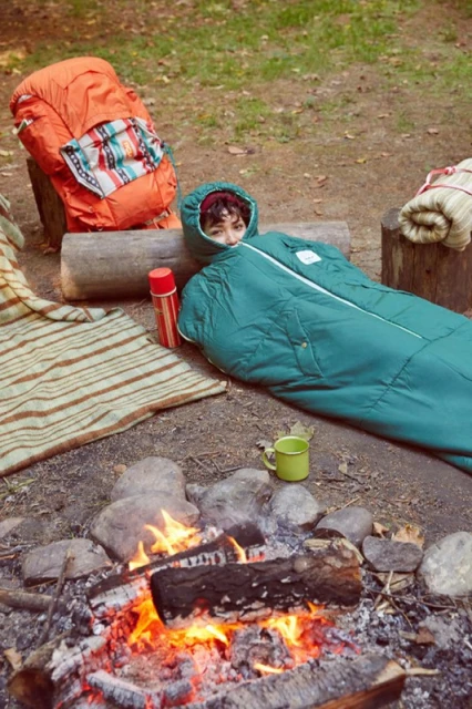 Young woman curled in a green sleeping bag next to a campfire in the forest with an orange camping backpack and other gear nearby.