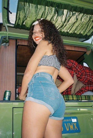 Woman poses beside vintage van in blue jean shorts, black-and-white gingham top, large hoop earrings, and a white headband, smiling outdoors.