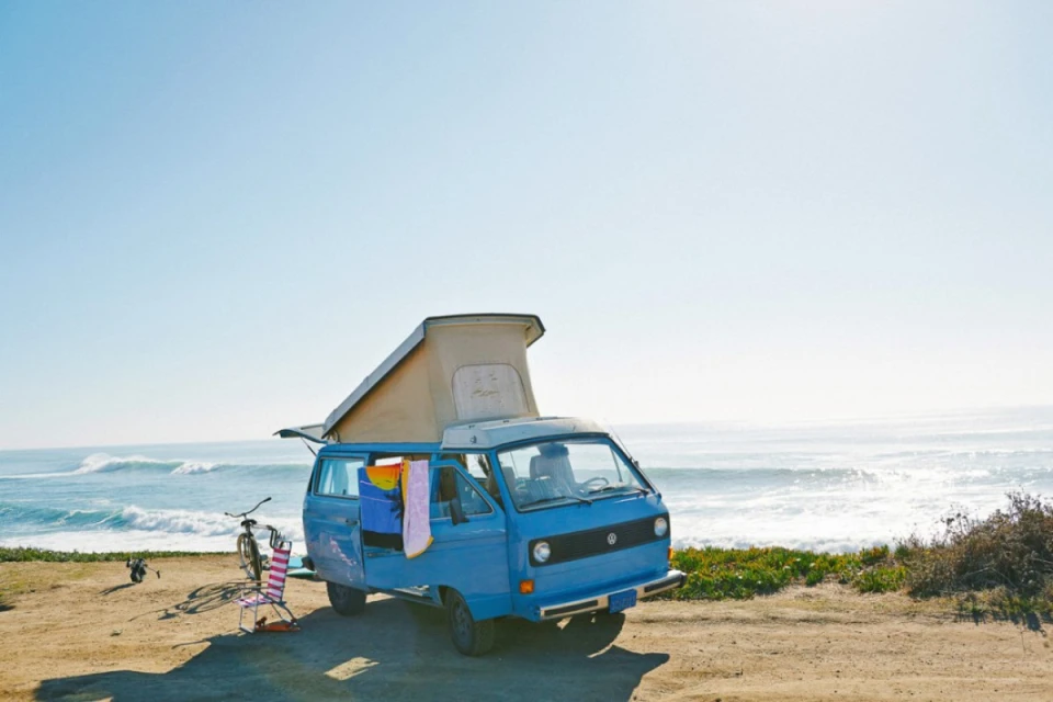 Blue Volkswagen camper van parked on a sandy beach with surf, blue sky, colorful beach chair, and bicycle in the background.