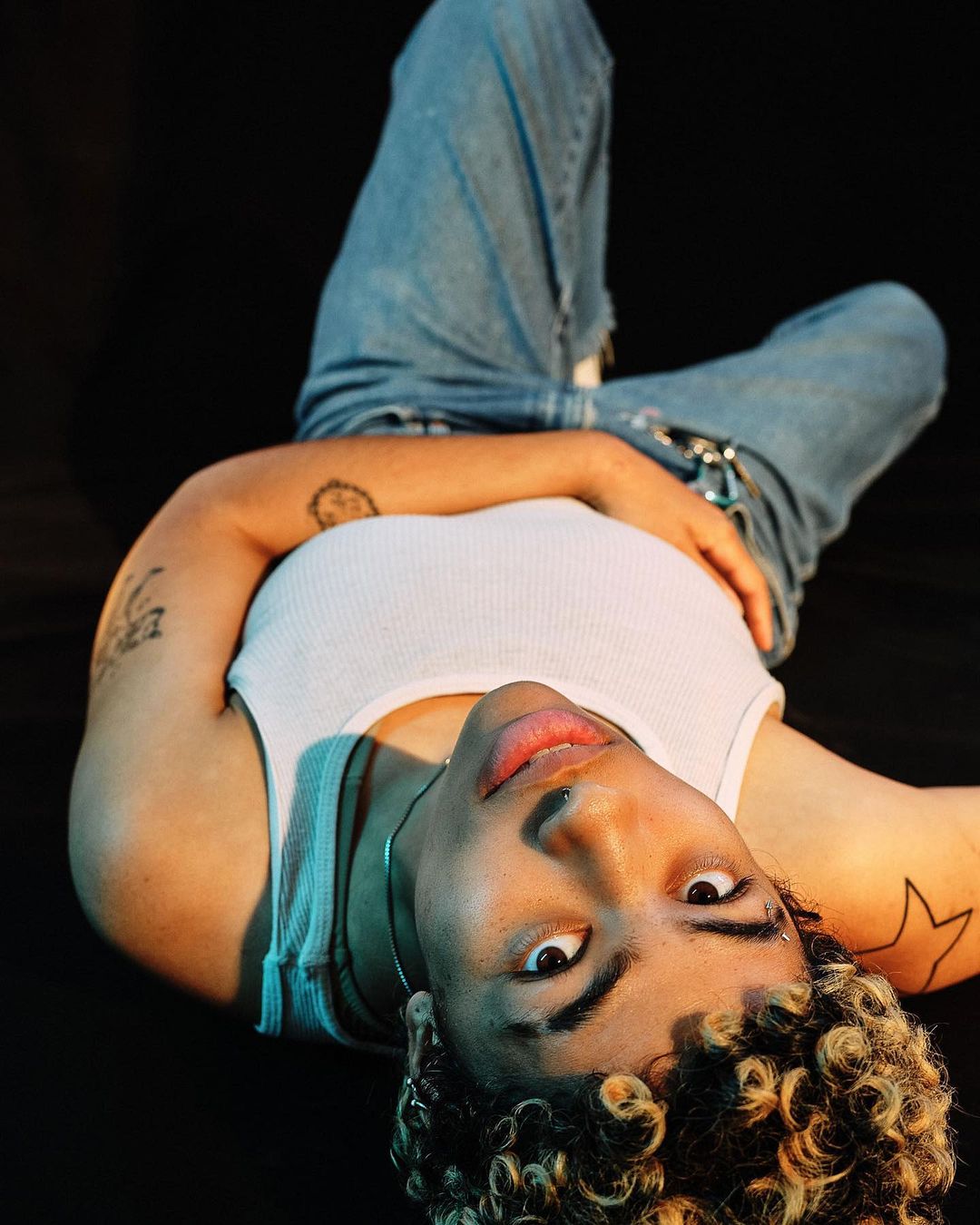 A young adult woman with curly blonde hair lies on a black surface, wearing a white tank top and light blue jeans, looking at the camera.