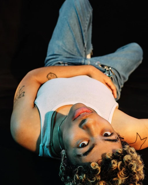 A young adult woman with curly blonde hair lies on a black surface, wearing a white tank top and light blue jeans, looking at the camera.
